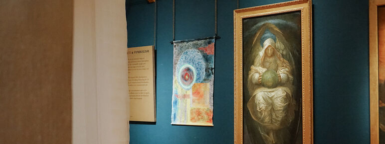 Photo inside the Historic Galleries at Watts Gallery. There is an abstract colourful artwork hung on the wall made from canvas. It is displayed beside paintings by G F Watts