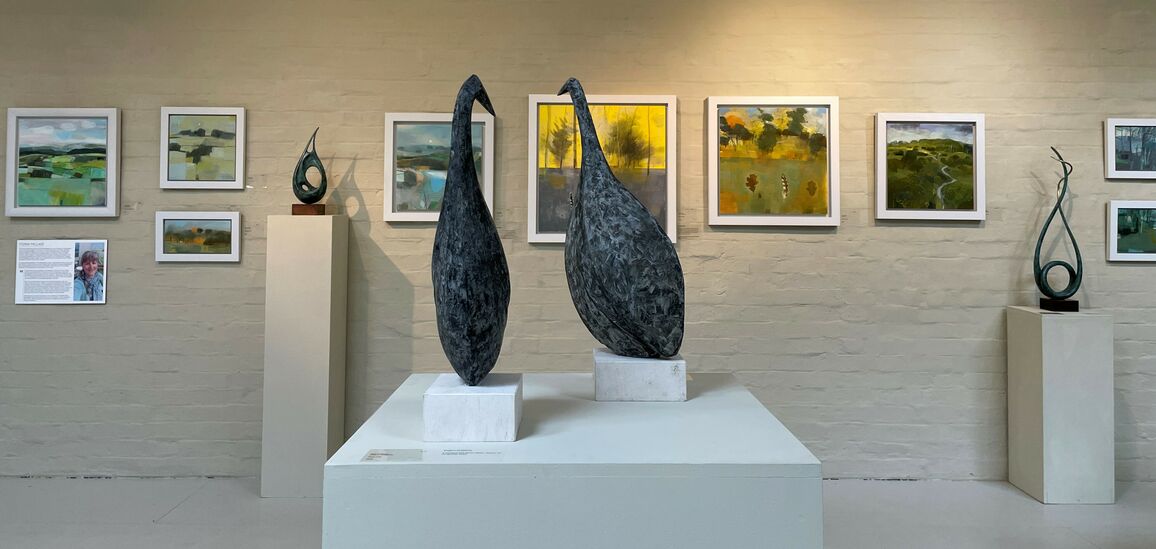 Two sculptures of guinea fowl in the foreground with framed landscape paintings on the wall in the background alongside two more small abstract bronze sculptures