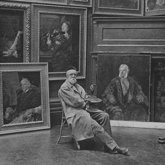 G F Watts posed on a chair in front of a large collection of portrait paintings