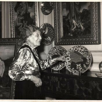 Black and white photo of a woman gesturing to a plate by William De Morgan placed on a mantelpiece