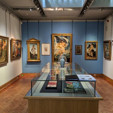 A view of the De Morgan Gallery looking towards Evelyn De Morgan's paintings, with a glass display case containing works by William De Morgan in the centre of the room