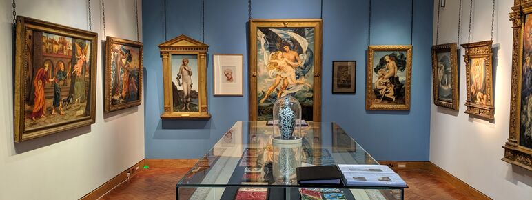 A view of the De Morgan Gallery looking towards Evelyn De Morgan's paintings, with a glass display case containing works by William De Morgan in the centre of the room