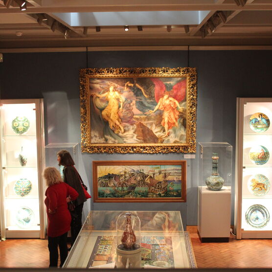 An old layout of the De Morgan Gallery with glass cabinets holding works by William De Morgan surrounded by oil paintings by Evelyn De Morgan hung on the walls