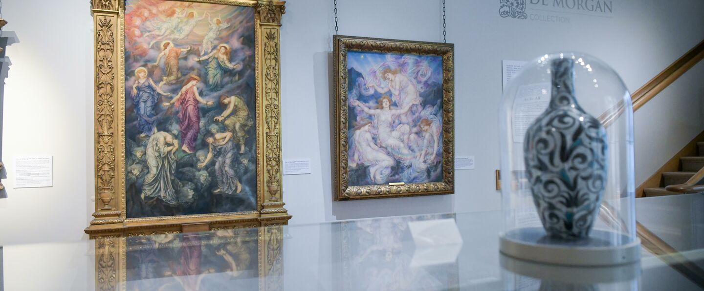 A floral vase encased in a glass display case with two colourful oil paintings in gold frames in the background