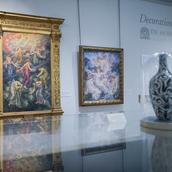 A floral vase encased in a glass display case with two colourful oil paintings in gold frames in the background