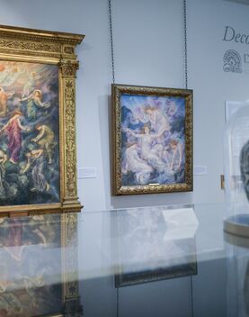 A floral vase encased in a glass display case with two colourful oil paintings in gold frames in the background
