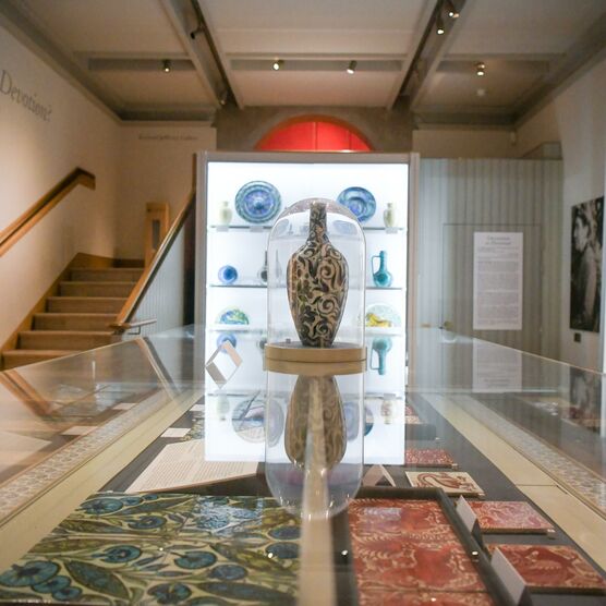 The De Morgan Gallery. Cabinets of plates and vases in the background, in the foreground, a large floral vase is encases in a glass cabinet on top of another cabinet containing terracotta tiles.