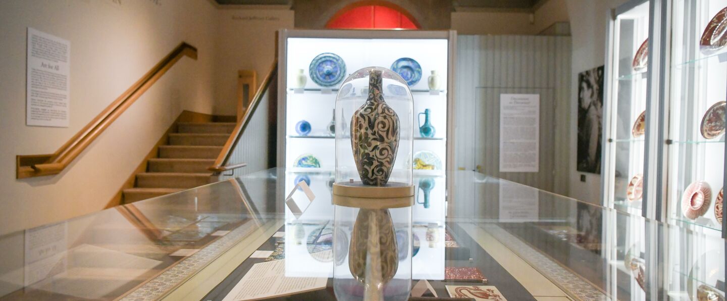 The De Morgan Gallery. Cabinets of plates and vases in the background, in the foreground, a large floral vase is encases in a glass cabinet on top of another cabinet containing terracotta tiles.