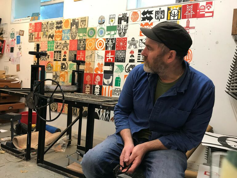 Photograph of a man sitting on a stool in his art studio. There are various prints hung on the wall behind him.