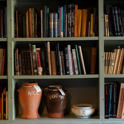 An image of Watts Gallery's archive room. There is sage green selving units full of different coloured books. In shelving at the bottom is two large ceramic pots. One orange rice pot and one brown sugar pot.