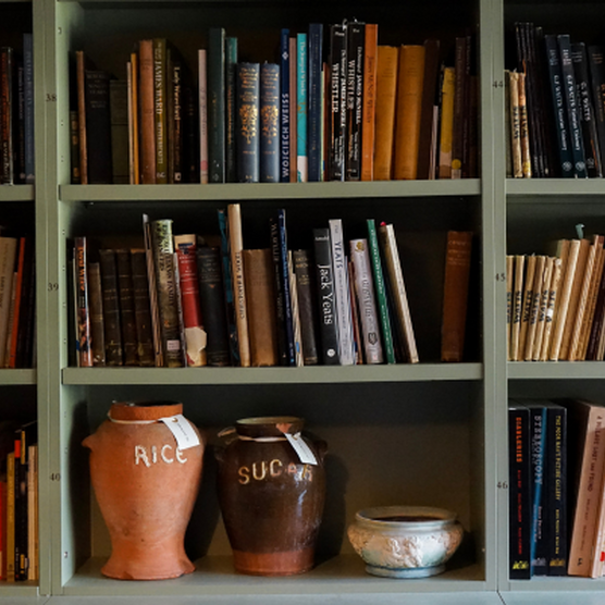 An image of Watts Gallery's archive room. There is sage green selving units full of different coloured books. In shelving at the bottom is two large ceramic pots. One orange rice pot and one brown sugar pot.
