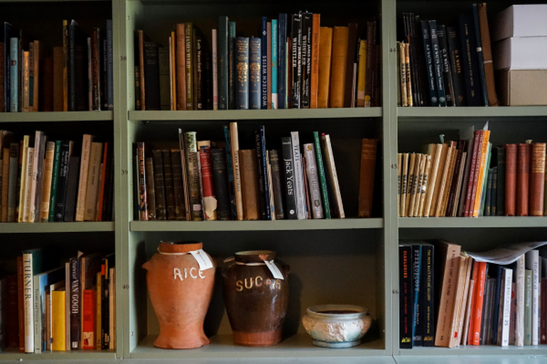 An image of Watts Gallery's archive room. There is sage green selving units full of different coloured books. In shelving at the bottom is two large ceramic pots. One orange rice pot and one brown sugar pot.