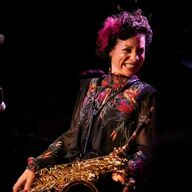 A photograph of a women smiling with brown and pink hair wearing a black dress holding a saxophone