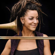A photograph of a women smiling with brown hair tied up and holding a trumpet