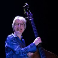 A photograph of a women smiling wearing glasses and a blue shirt looking at the camera holding a cello