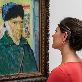 A photograph of a women wearing a headset looking at a Van Gogh painting hung on the wall