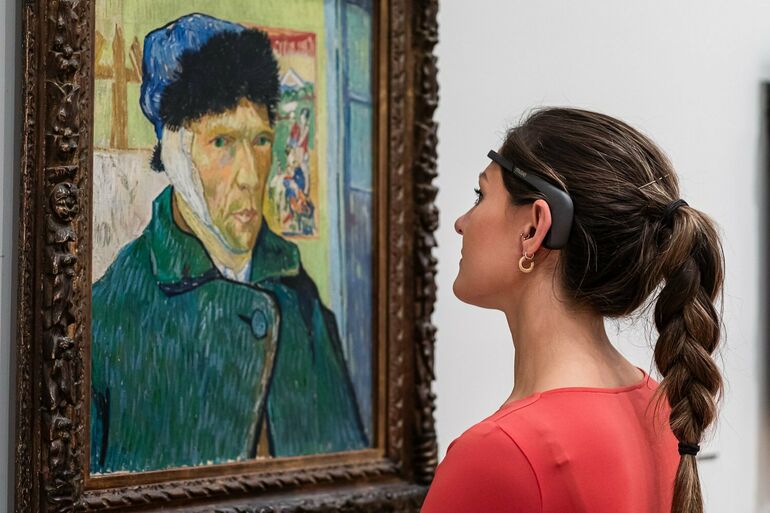 A photograph of a women wearing a headset looking at a Van Gogh painting hung on the wall