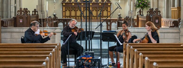 Two men and a woman playing violin's whilst reading music and another woman playing the cello whilst sat inside a church