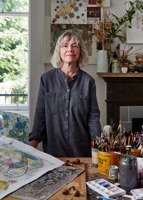 Portrait photograph of Angie Lewin standing behind a table which has her prints and paintbrushes on. Her hair is grey and cut in a bob with a fringe, and she is wearing a charcoal coloured collarless shirt.