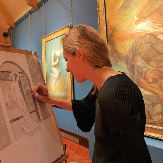 A woman with blonde hair sketches the Watts Historic Gallery at an easel