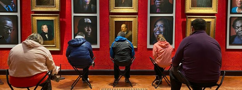 5 people sat on stools in the Historic Gallery facing the Faces of Fame installation, they are hunched over drawing and their backs are to us