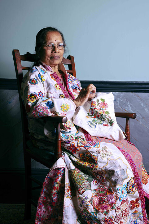 An elderly woman wearing an embroidered gown, sat in a high-backed wooden chair