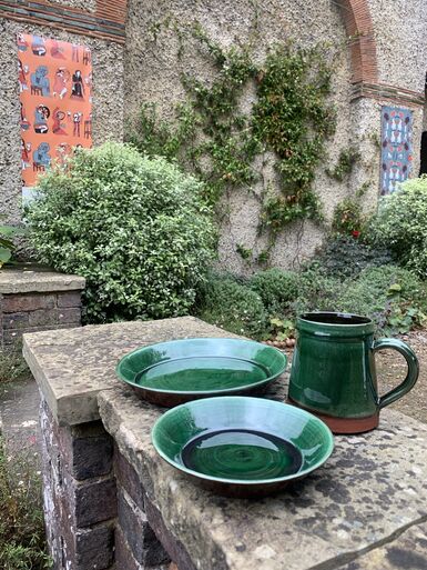 Two green plates and a green mug laid out on a low wall next to Watts Gallery