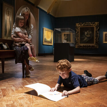A boy lies on the gallery floor reading while his mother sits on a bench watching him, holding another young child.