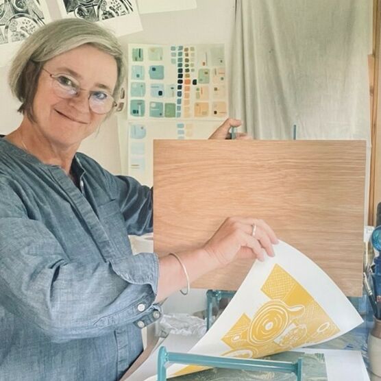 Photo of a woman with short grey hair pulling a print from a printing block. She is smiling at the camera.