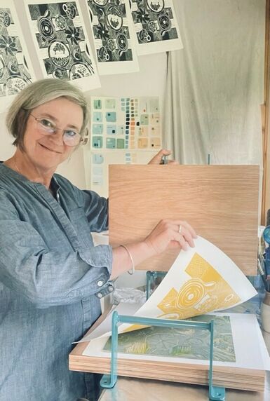 Photo of a woman with short grey hair pulling a print from a printing block. She is smiling at the camera.