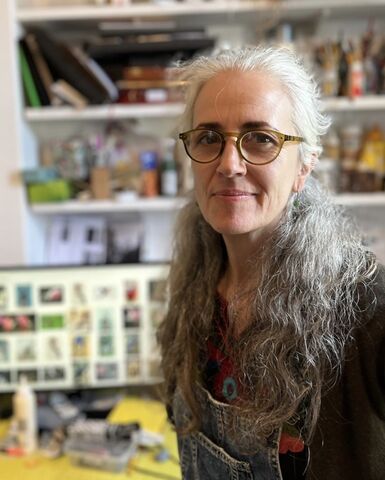 Photo of a woman with long grey curly hair and round glasses. She is facing the camera and smiling