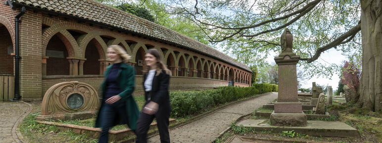 Two visitors walk along a stone path in the cemetery. There are terracotta archways in the background.