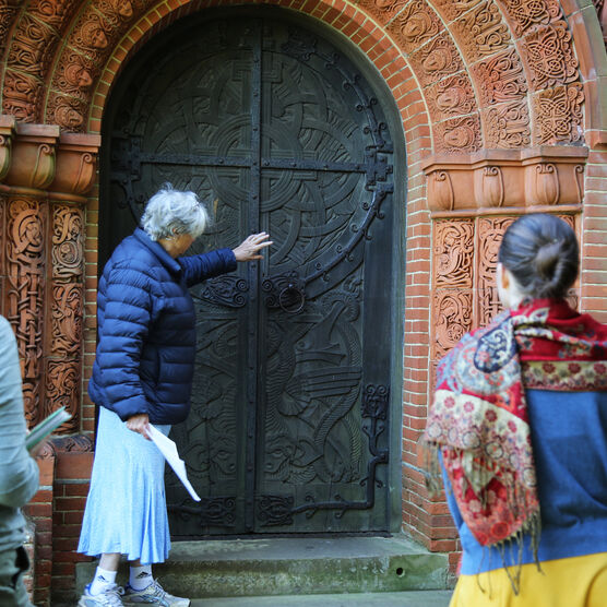 A group of four gather around the highly-decorated door of Watts Chapel. The tour guide points at the door.