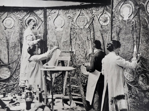 A black and white photograph of Mary with three villagers decorating a highly-decorated panel wall.