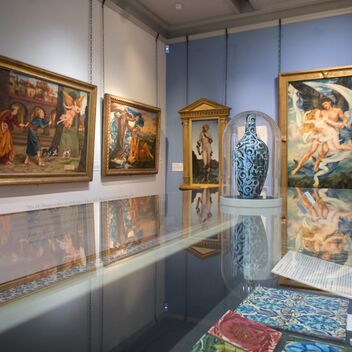 A white and blue-walled gallery with gold framed paintings and tiles in a glass case