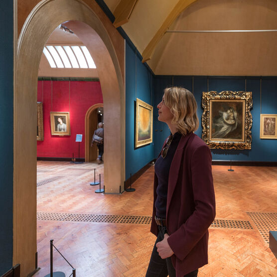 A visitor views a gold-framed painting in the Historic Galleries, in the background are more paintings on a blue wall and the stone statue, Aurora.
