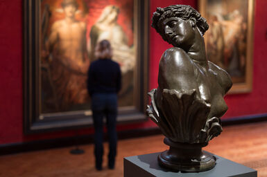 A bronze sculpture of Clytie, a bust turning her head in profile. In the background a visitor views paintings on the red wall.