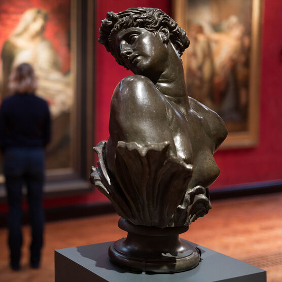 A bronze sculpture of Clytie, a bust turning her head in profile. In the background a visitor views paintings on the red wall.