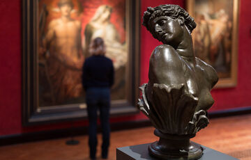 A bronze sculpture of Clytie, a bust turning her head in profile. In the background a visitor views paintings on the red wall.