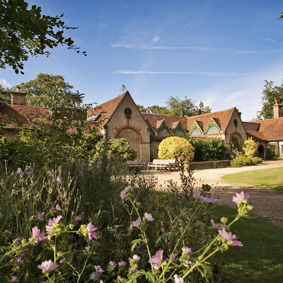 Outside Watts Gallery, the sky is blue, the building is beige with a triangular roof, there are flowers in the foreground.
