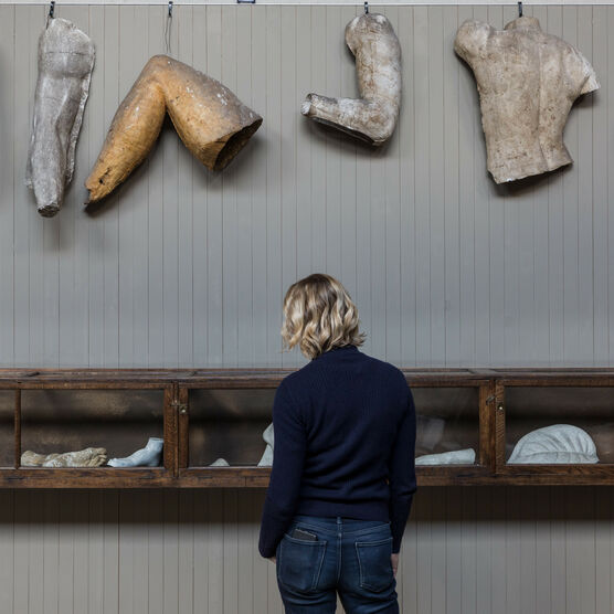 The Sculpture Gallery at Watts Gallery, the wall is grey with plaster casts of various limbs hanging in a row, central to the image is a woman that has her back to the camera. She is looking at a display case filled with various sculptural objects.