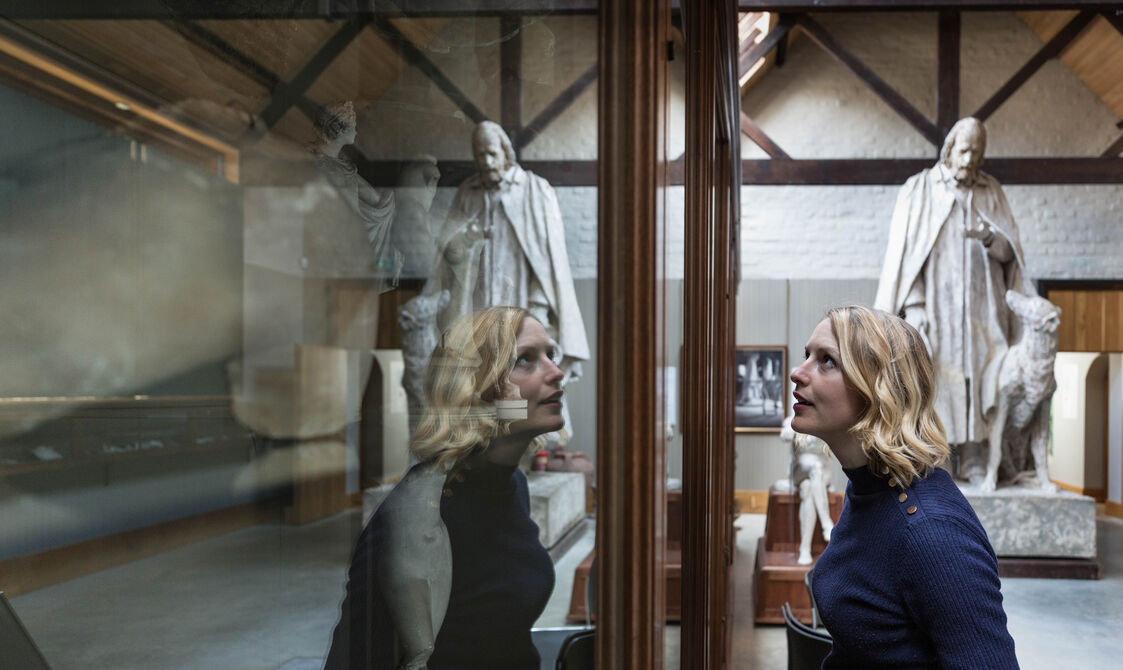 A women views sculptural objects in a case in the Sculpture Gallery. In the background is the plaster sculpture of Alfred Lord Tennyson and his dog.