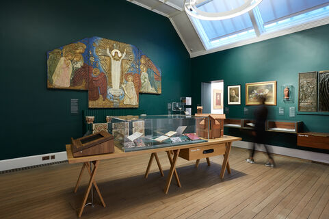 A green gallery with artworks on the wall and a wooden table with a glass case, filled with objects