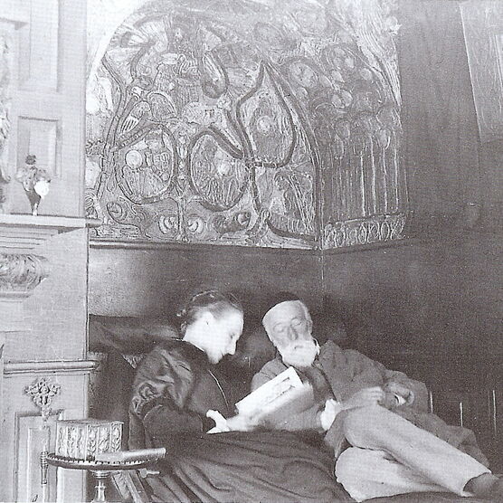 Archive black and white photo of George and Mary, Mary reads to George underneath a decorated arch way