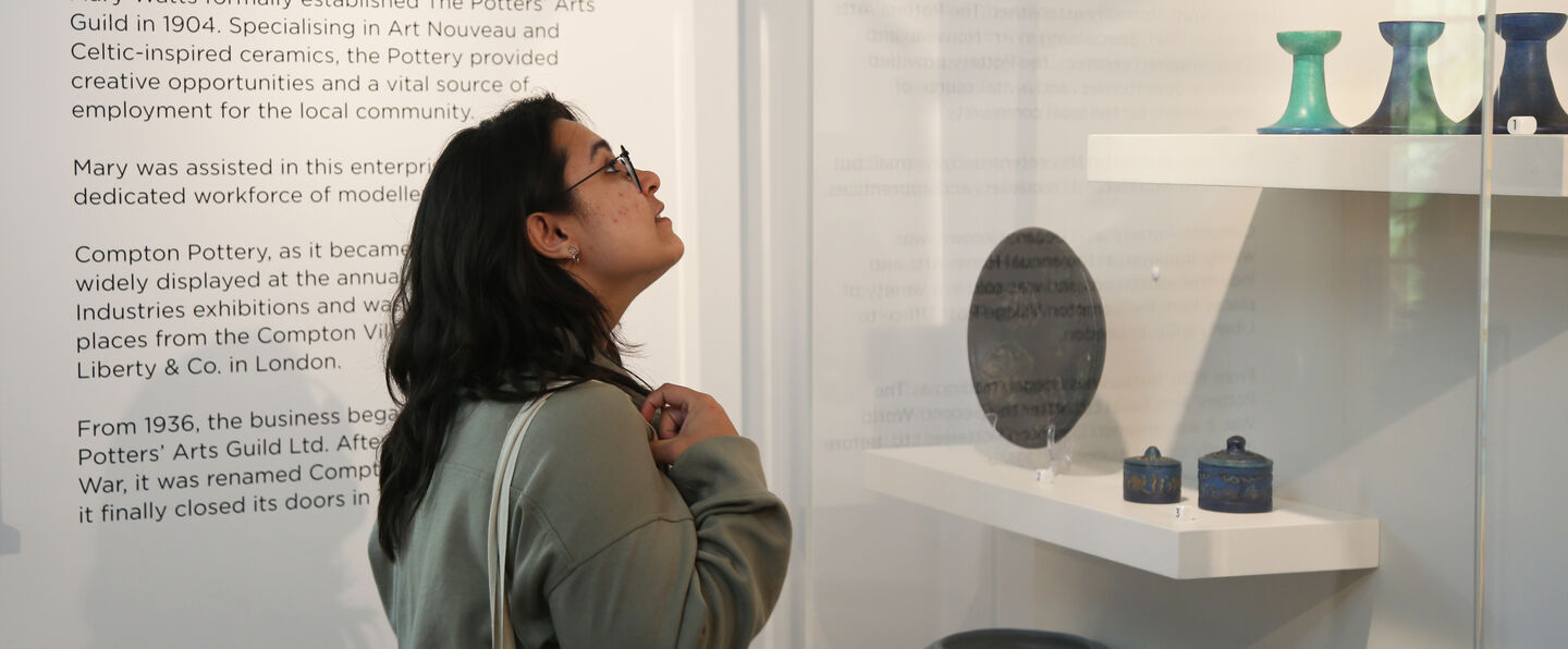 A young woman looks at examples of Compton pottery in a glass display case
