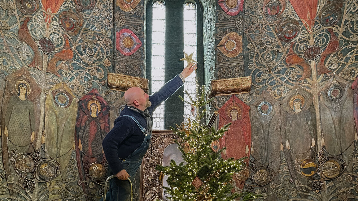 Photograph of a man standing on a ladder and placing a star on the top of a Christmas tree inside Watts Chapel. The walls of the chapel are decorated with angels made from painted gesso.