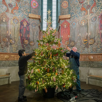 Photograph of a Christmas tree in the middle of Watts Cemetery Chapel. The walls of the chapel are decorated with angels made from painted gesso. A male and a female are decorating the tree.