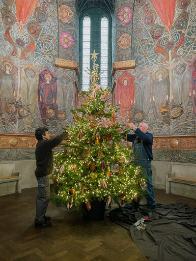 Photograph of a Christmas tree in the middle of Watts Cemetery Chapel. The walls of the chapel are decorated with angels made from painted gesso. A male and a female are decorating the tree.