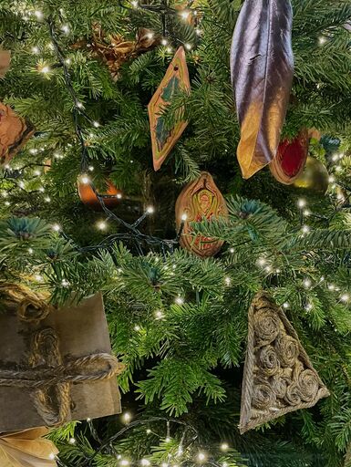 Close-up photograph of a Christmas tree. Decorating the tree are fairy lights, hand painted terracotta decorations and other decorations made out of paper and card.